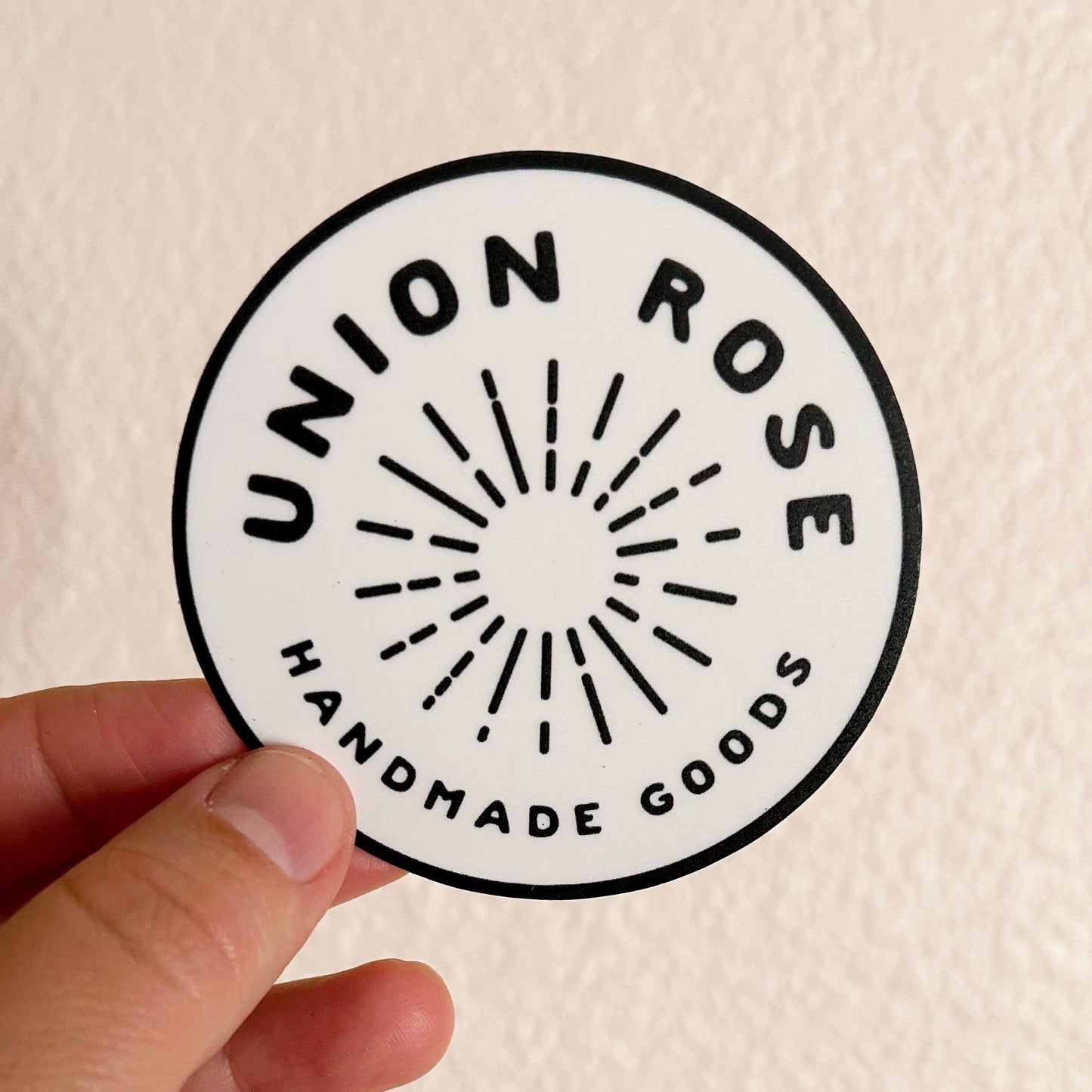 Hand holding a round sticker with 'Union Rose Handmade Goods' logo on a beige background
