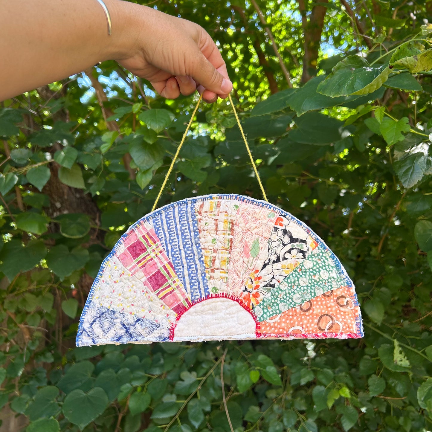 Colorful patchwork fan-shaped ornament held by a hand against a green leafy background