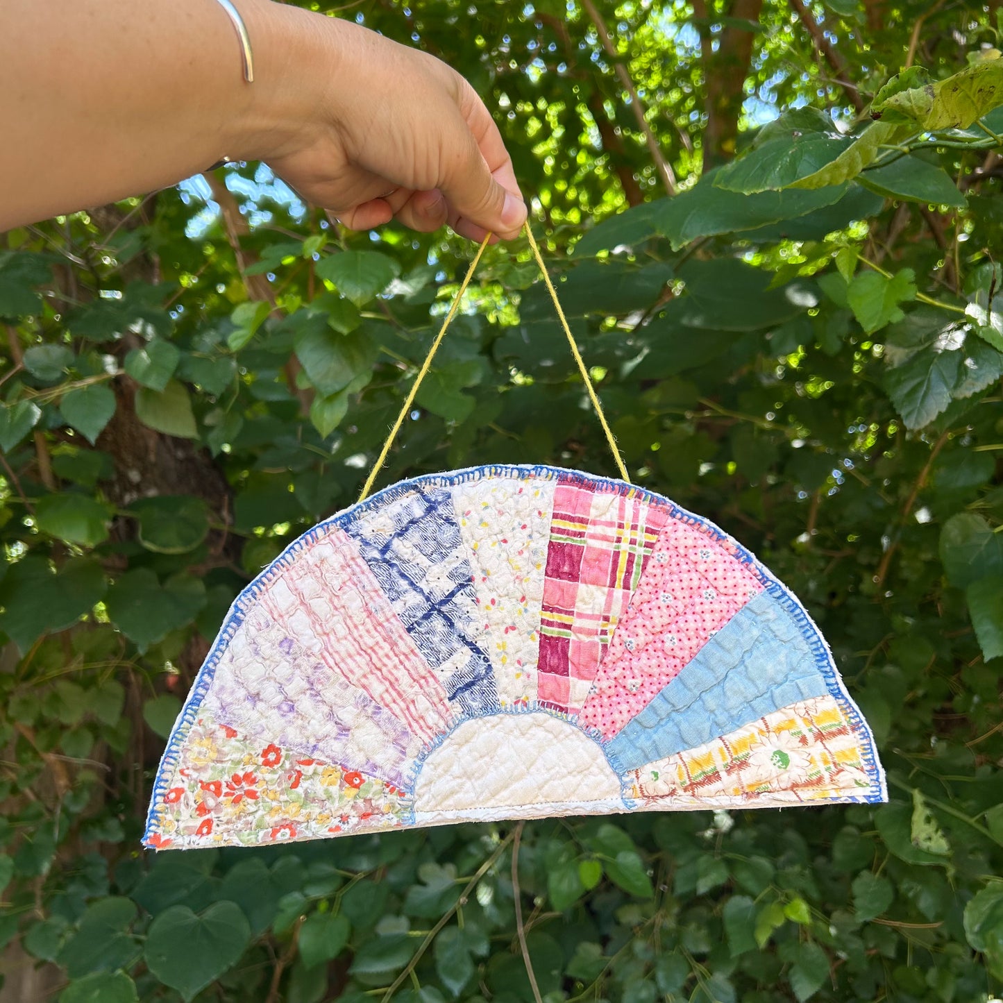 Handmade patchwork fan held by a hand against a green leafy background