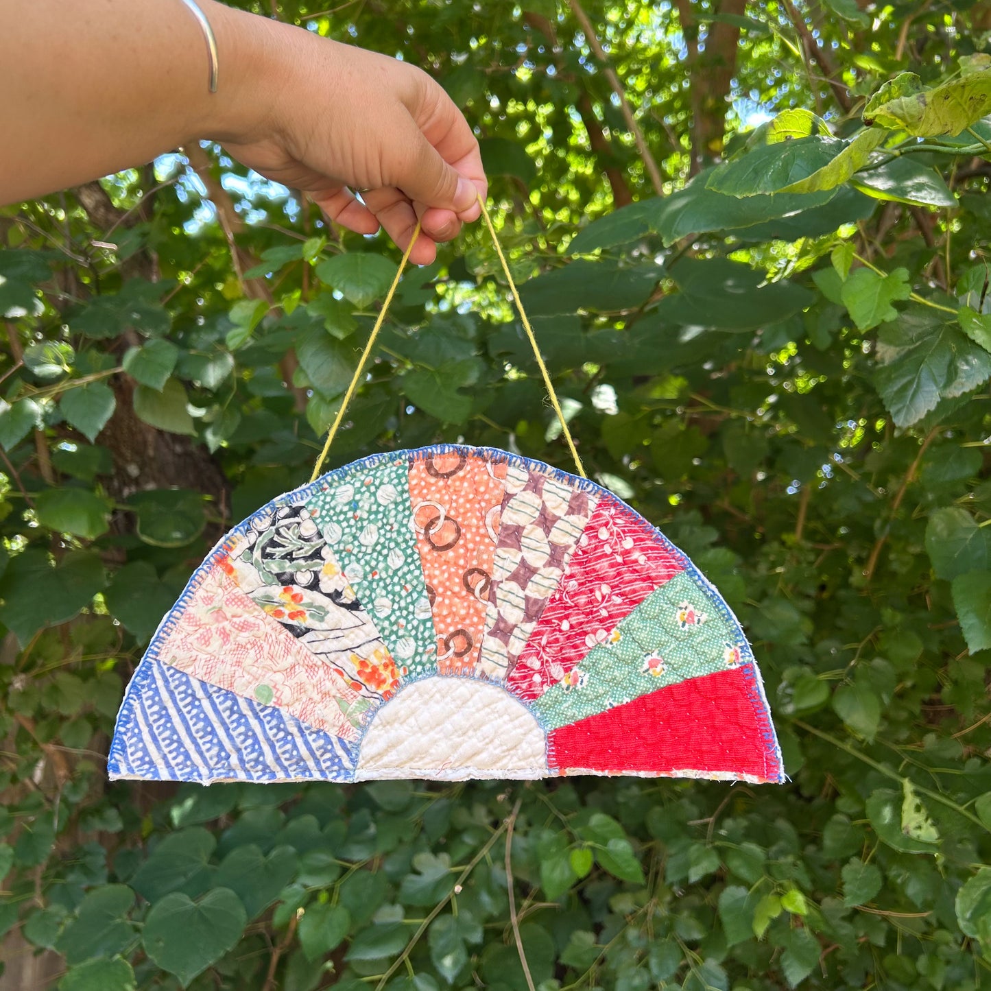 Handmade patchwork fan held by a hand against a green leafy background