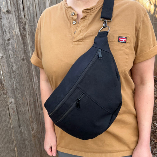 A person wearing a brown shirt is carrying an oversized black canvas crossbody bag with zipper pockets and a wider strap.